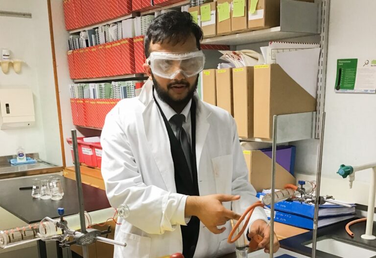 Male VI student in white laboratory coat in the Science room. He is using an adapted syringe to measure out acid and adding it to calcium carbonate