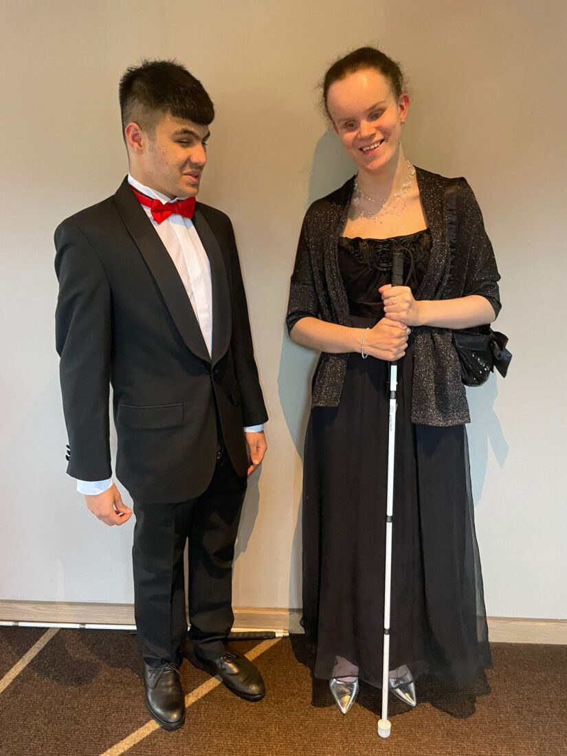 Mustafa is dressed in a black suit with red bow tie. Martha is in a black dress holding her cane. Both students are smiling at the camera