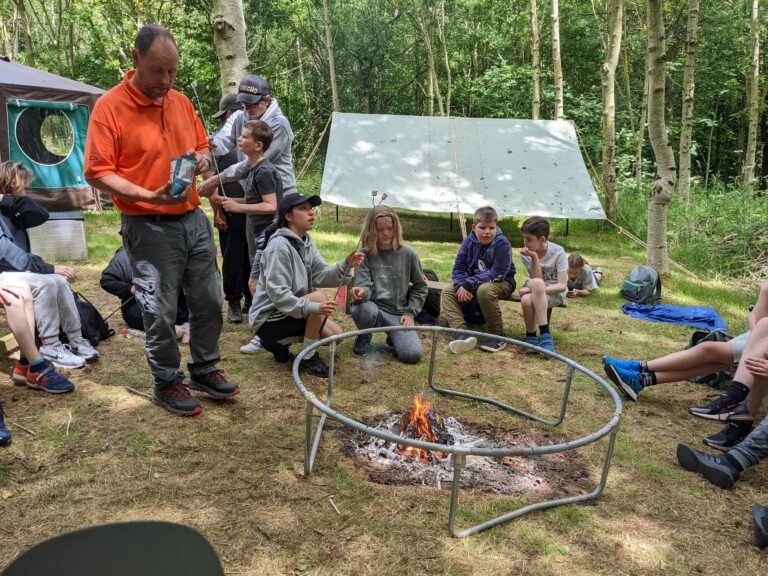 Students are sat around the camp fire, getting ready to toast some marshmallows