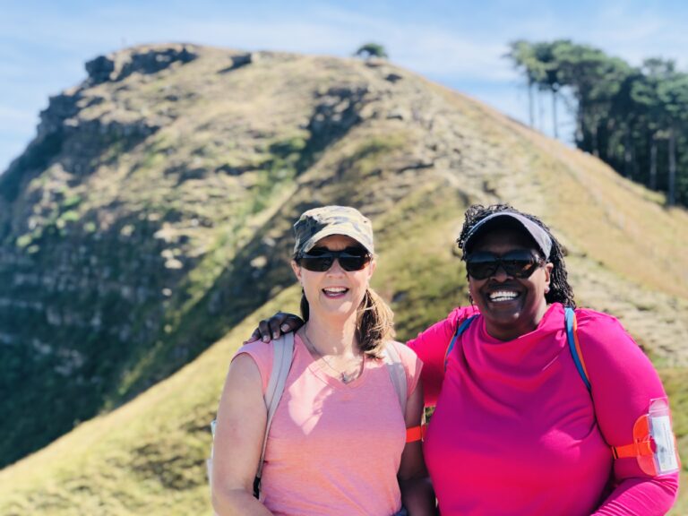 Image of Gigi and Sue. There is a Peak in the background they have have just climbed. The Sky is blue and the weather is sunny