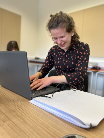 image of a student working on a laptop with printed braille pages to their left. The student is smiling