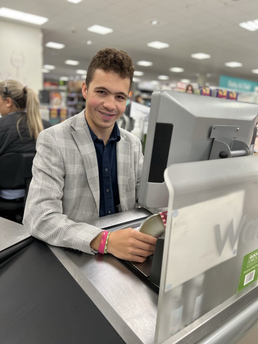 Student Thomas behind the till at Waitrose supermarketing, smiling at the camera