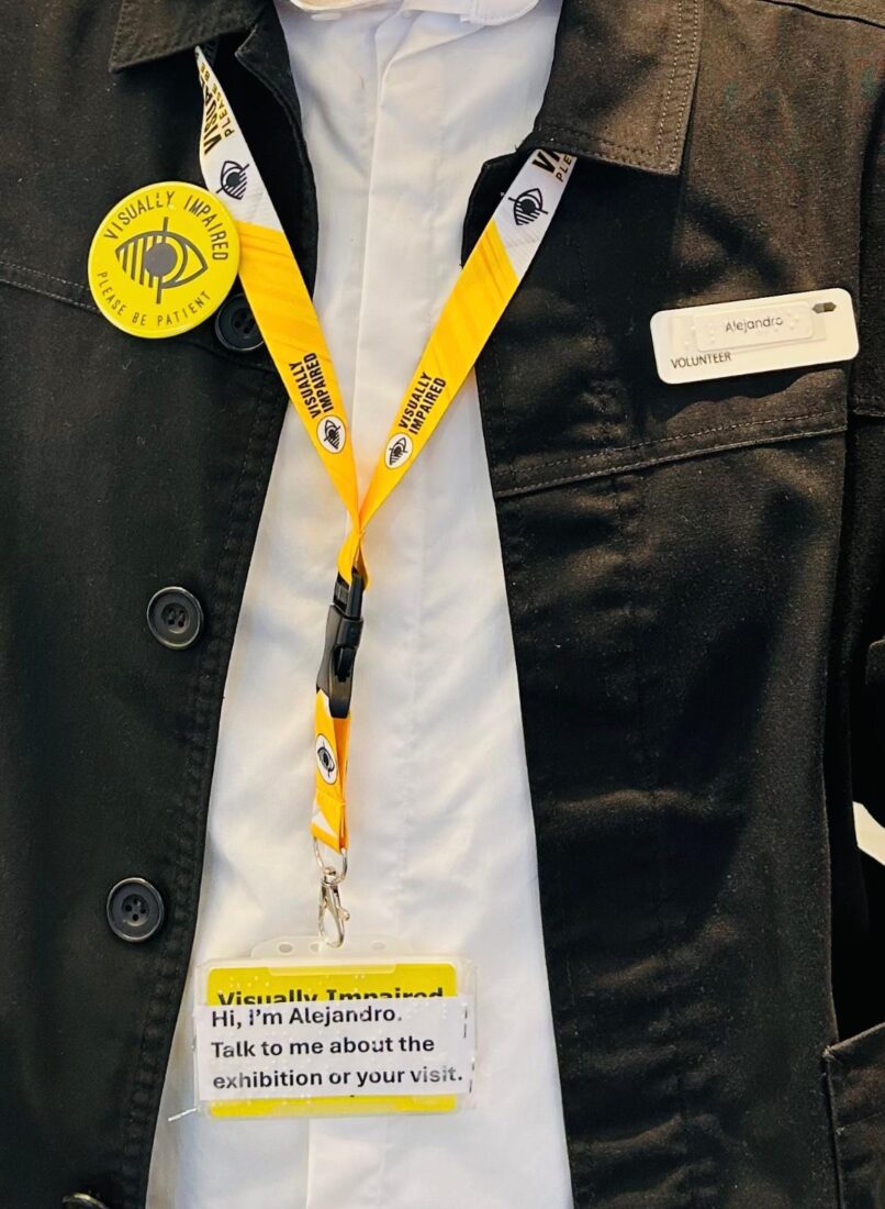 A close-up of Alejandro’s name tag and lanyard in Braille and print. He also has the yellow button on his jacket that has the universal blind/visually impaired symbol on it. Alejandro and his manager came up with the message on his lanyard.