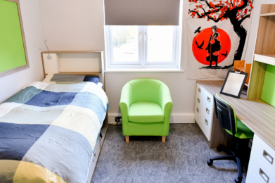 photo of a student's room, the bed is neatly made, there is a bright green tub chair, a clean and tidy desk and some artwork on the walls.