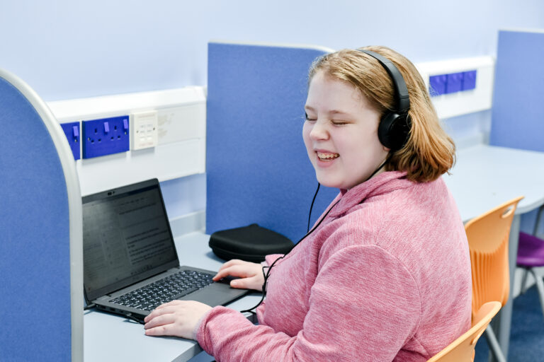 image of student smiling, wearing headphone whilst working at a laptop