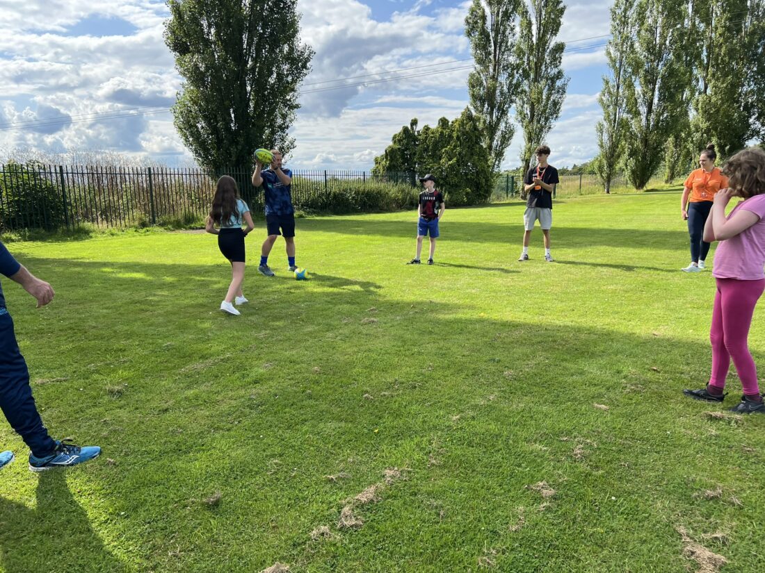 An image of a group of young people stood in a circle on a field passing a rugby ball