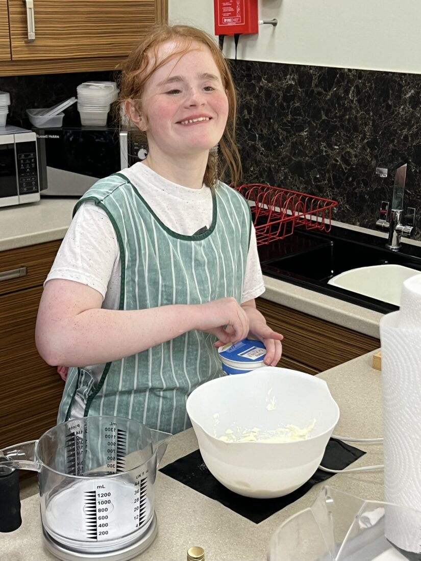 Image shows a young person smiling and wearing an apron doing some baking in a kitchen